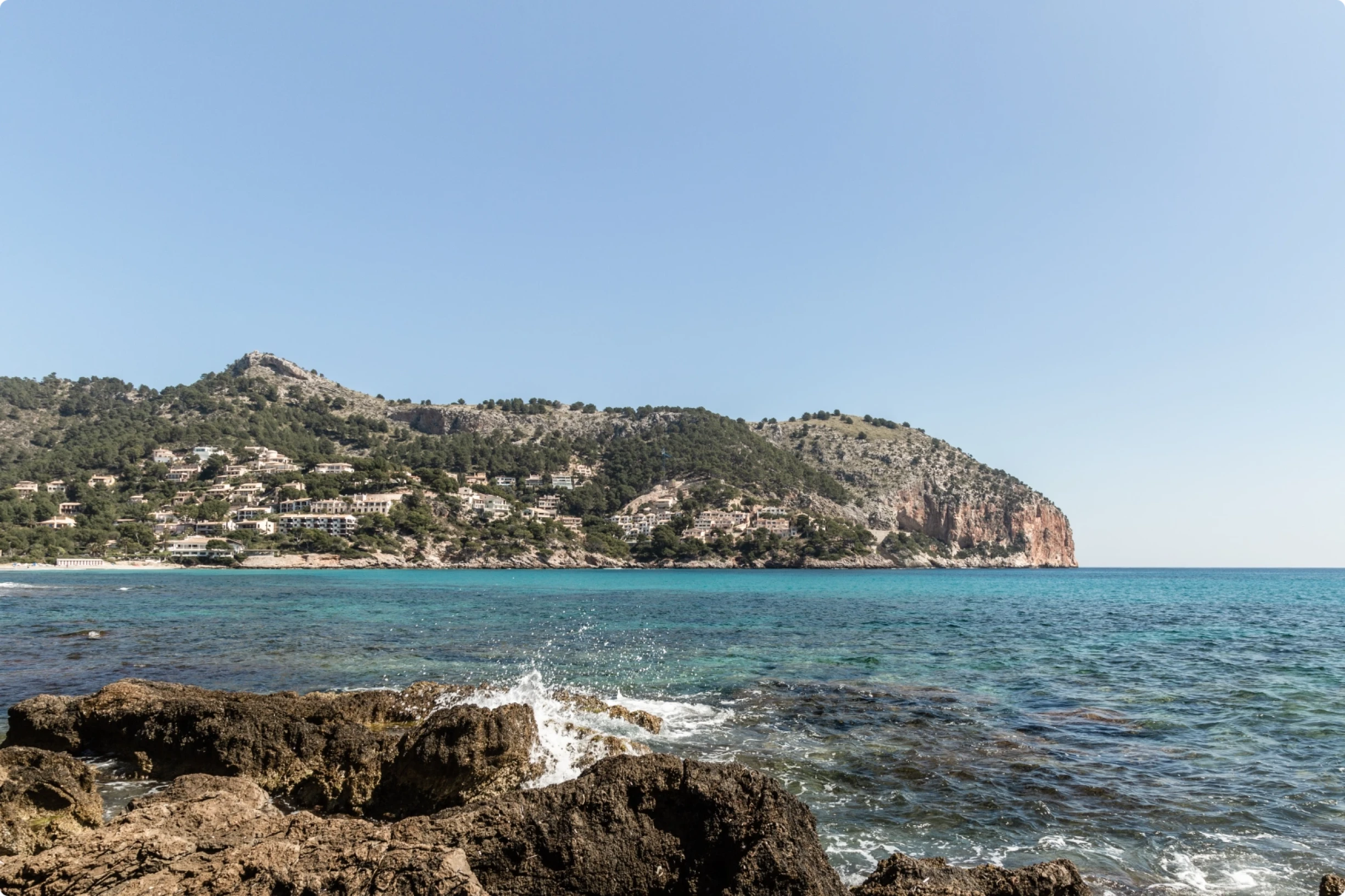 Rocky shoreline with waves crashing in the foreground and a coastal village nestled on a forested mountain.