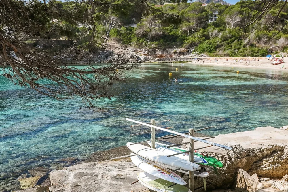 Paddleboards stacked on a rocky shore near clear turquoise water and a sunlit beach.