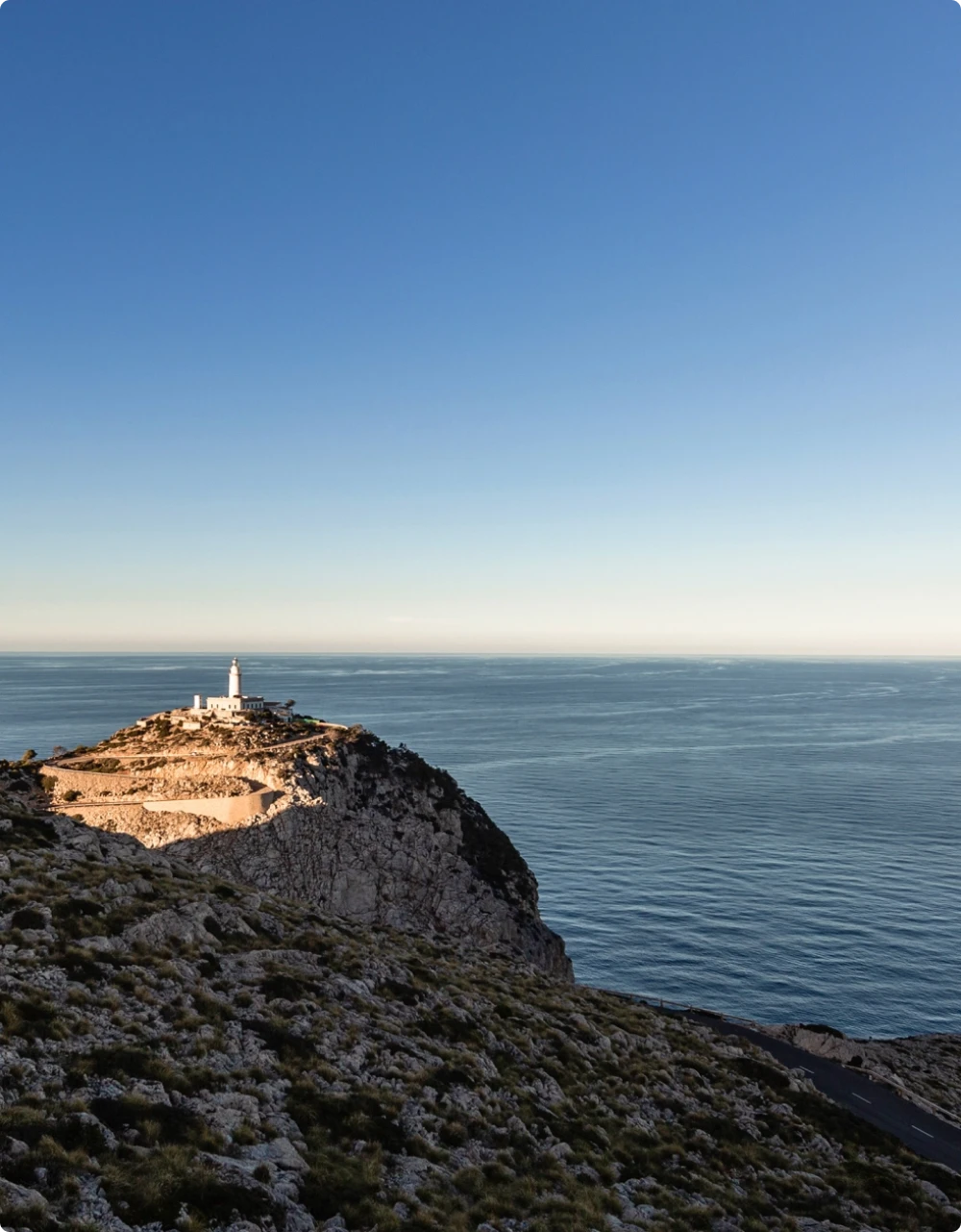 A white lighthouse perched on a steep, rocky cliff overlooking a vast, calm blue sea under a clear sky.