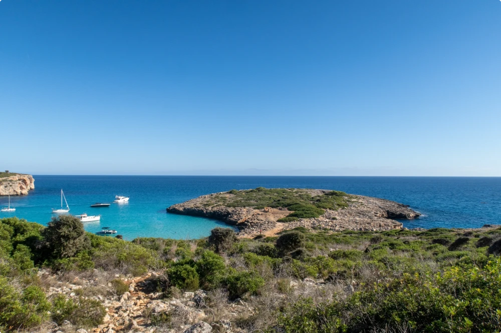 Coastal landscape with turquoise sea, a small rocky island, and boats anchored in a calm bay under a clear blue sky.