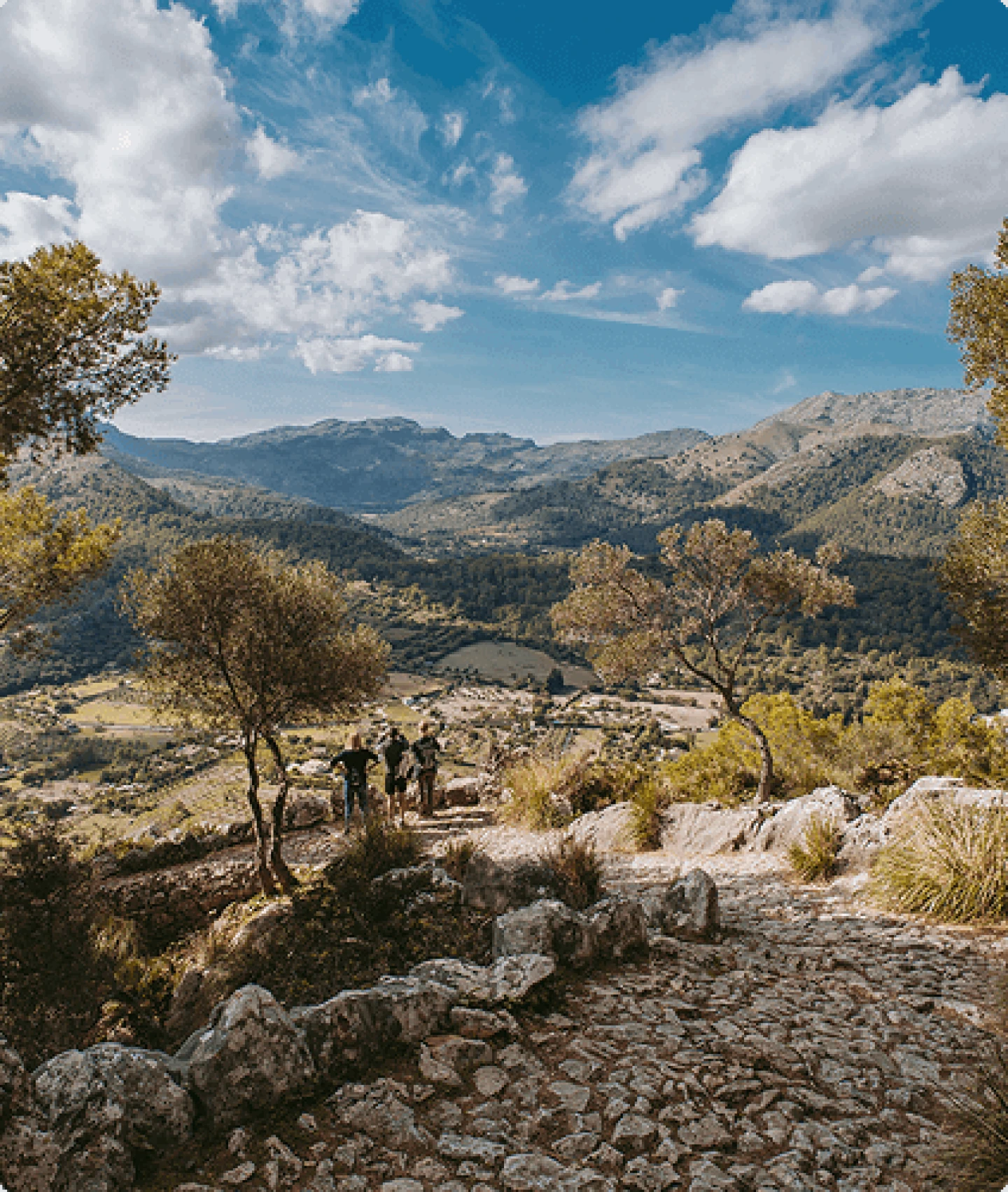 Hikers on a stone trail overlooking a lush green valley and mountains near Pollença