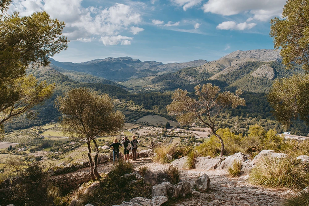 Three hikers on a mountain trail overlooking a lush valley and rugged peaks in Pollensa.
