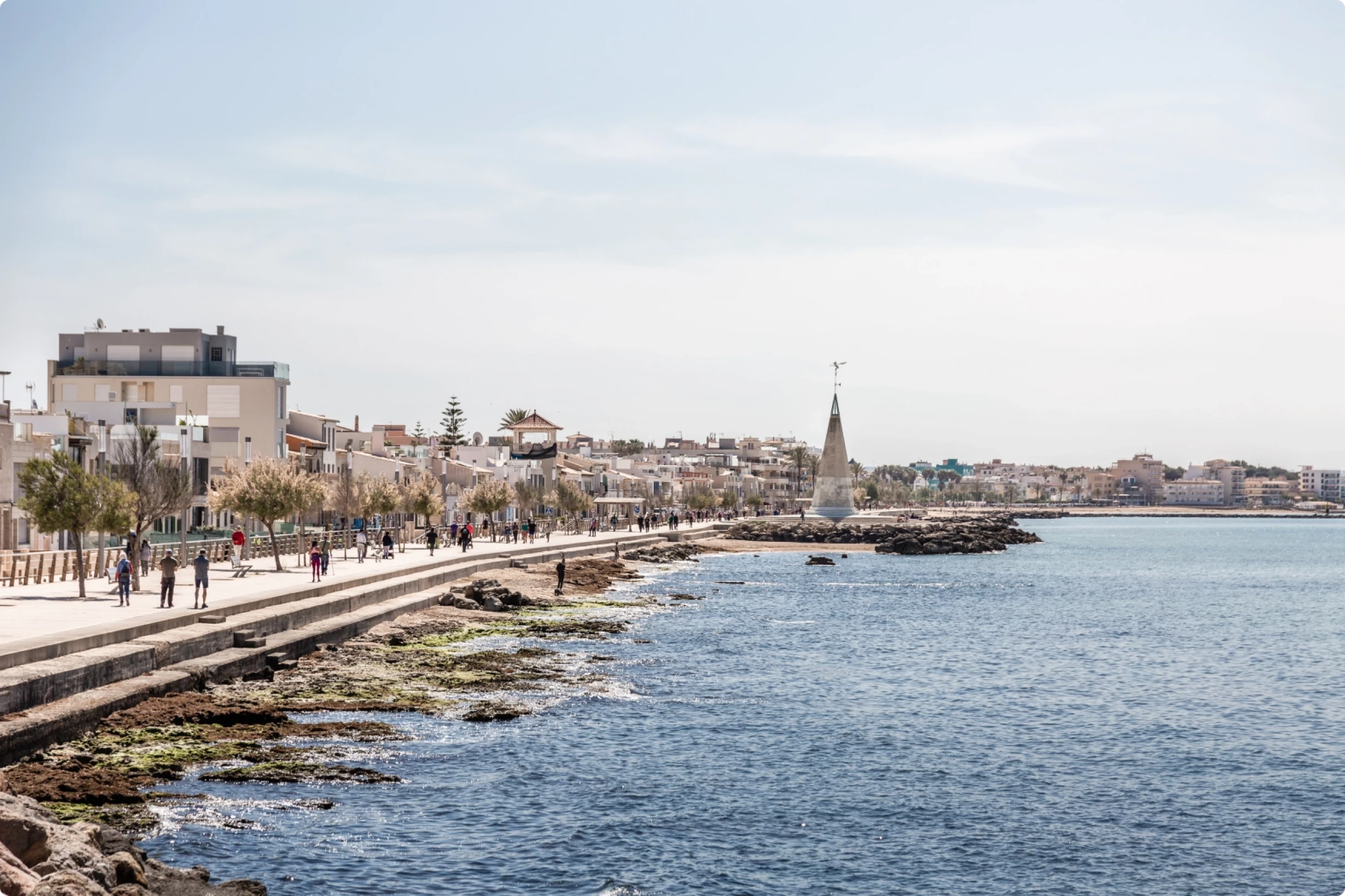Seafront promenade in Portixol, Mallorca, with people walking along the waterfront, coastal buildings, and calm blue sea under a bright sky.